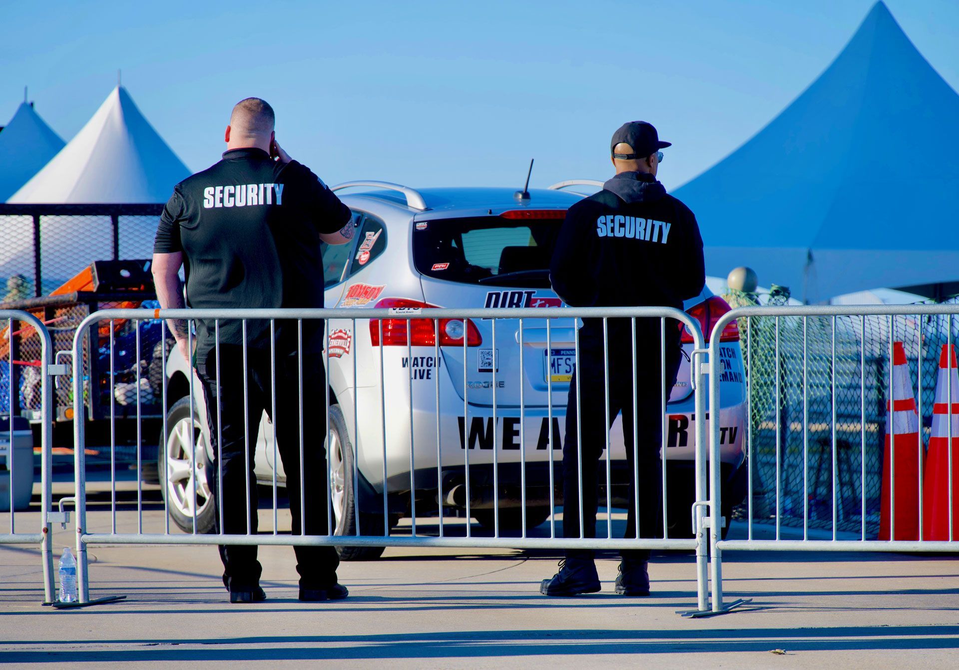 Two security guards stand by a white car behind metal barricades at an outdoor event.