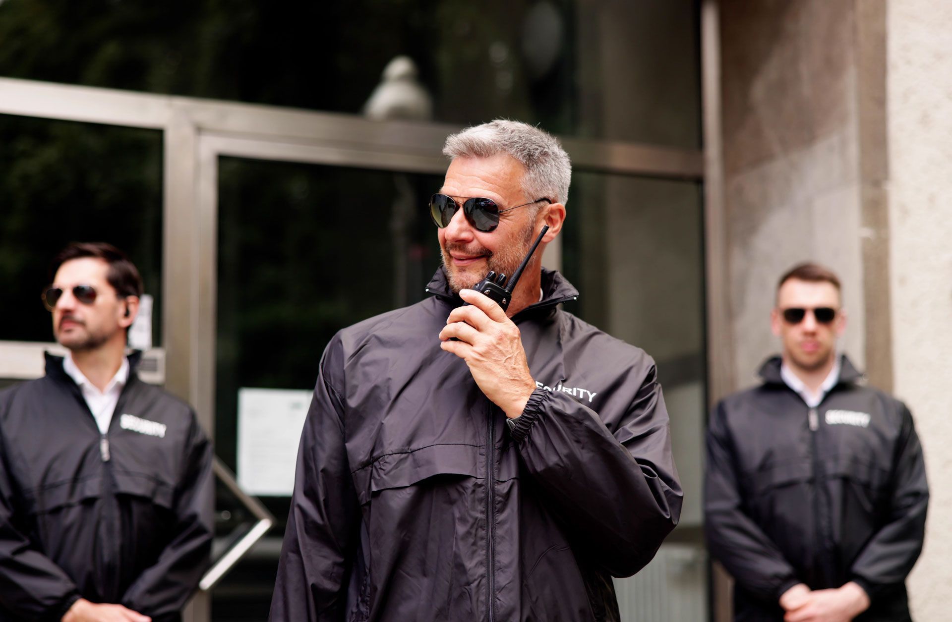 Three men in black jackets and sunglasses stand outside a building, with the center man speaking into a walkie-talkie.