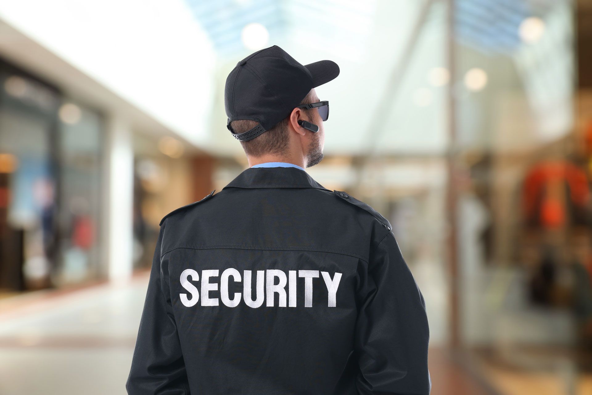 Security guard in black jacket and cap standing in a bright indoor corridor, seen from behind