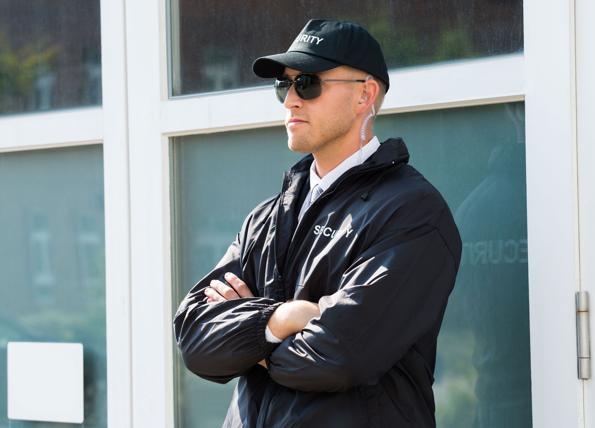 Man in black jacket and cap standing with arms crossed by a window