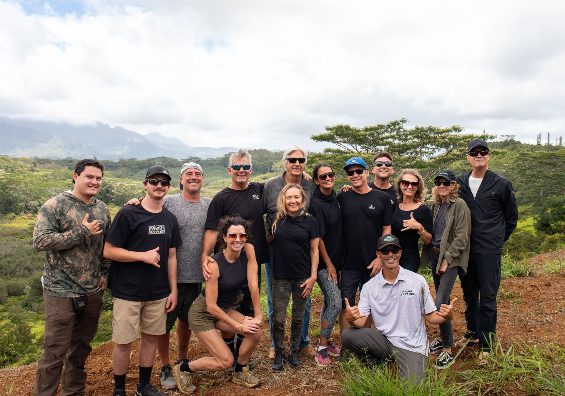 Group of people posing outdoors on a muddy hill with cloudy sky and greenery in the background.