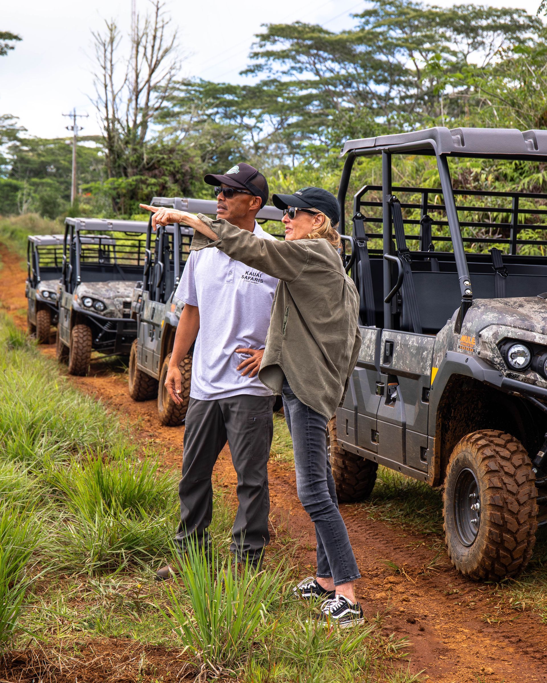 Two people stand by off-road vehicles on a muddy trail, one pointing ahead in a green landscape.