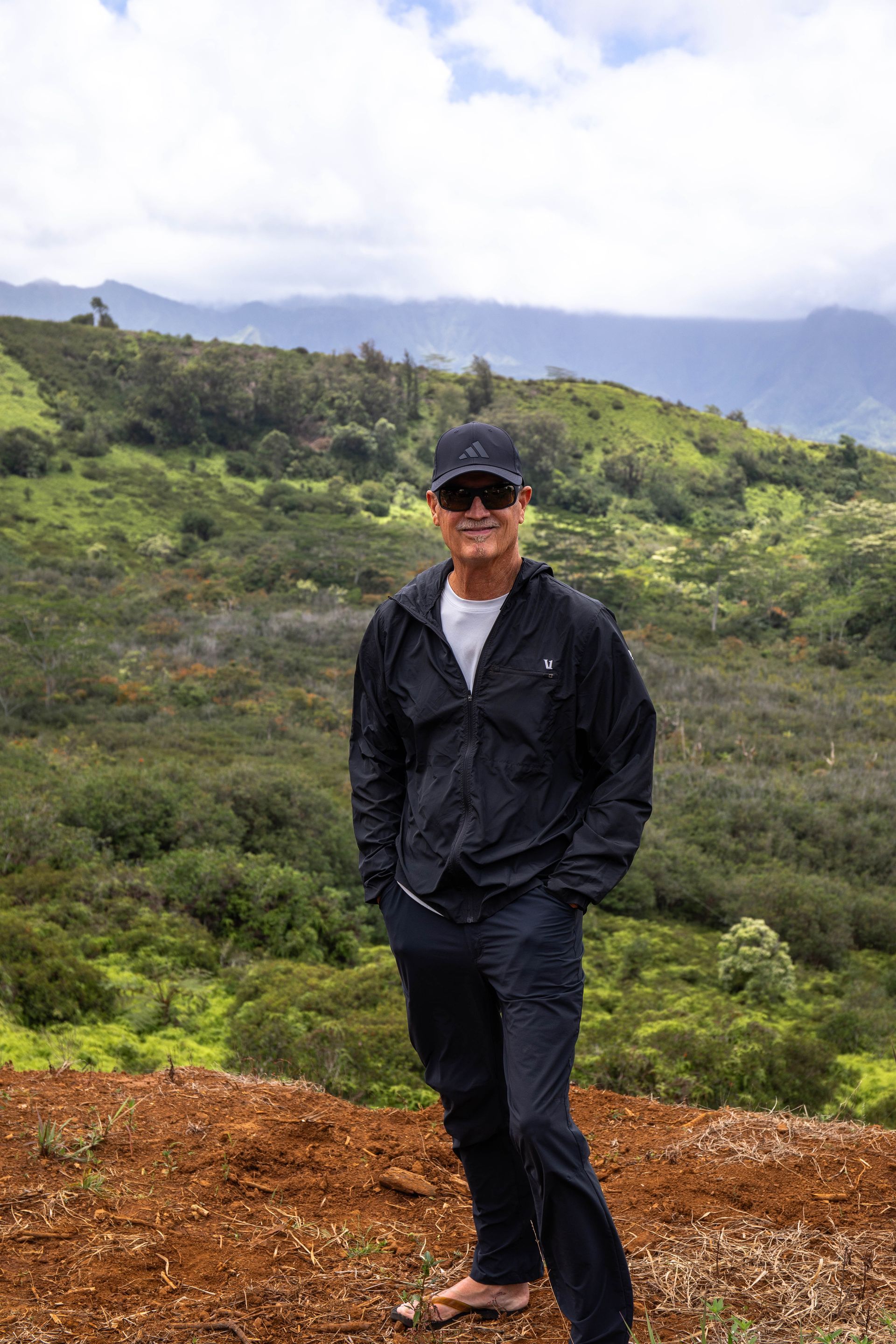 Person in black jacket and cap standing on a hillside with green mountains and cloudy sky in the background