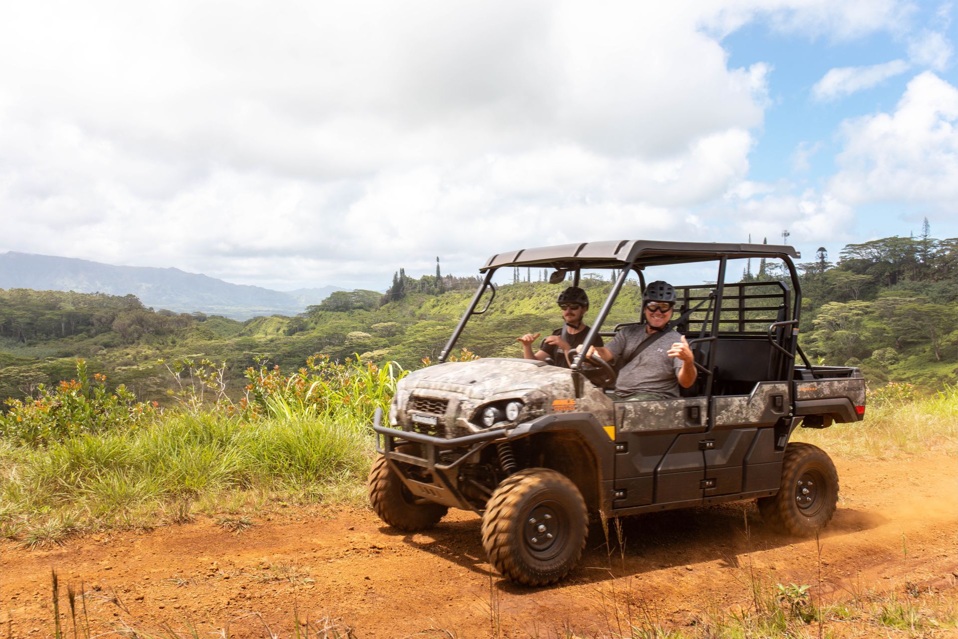 Two people ride in a black off-road buggy on a dirt trail in a grassy mountain landscape.