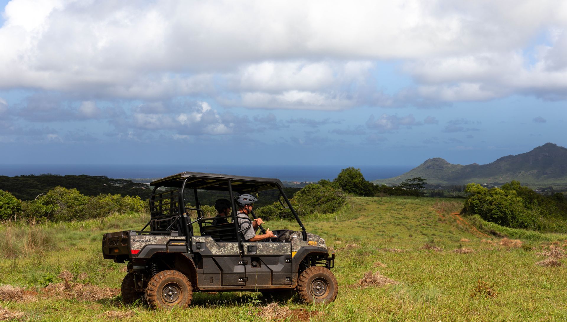 Off-road vehicle on grassy hillside overlooking ocean and mountains under cloudy sky