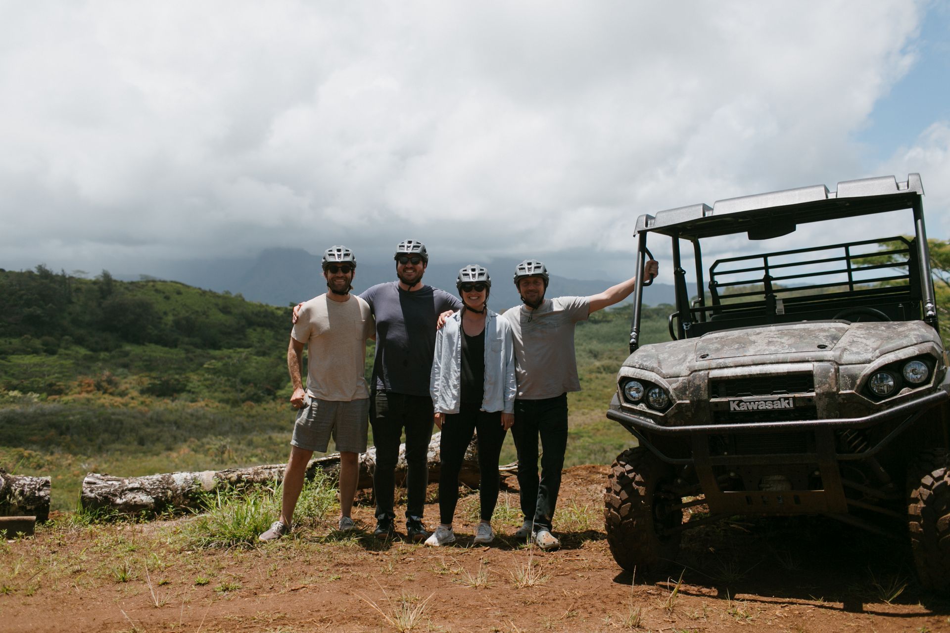 Four cyclists posing beside an off-road vehicle on a dirt road under cloudy skies.