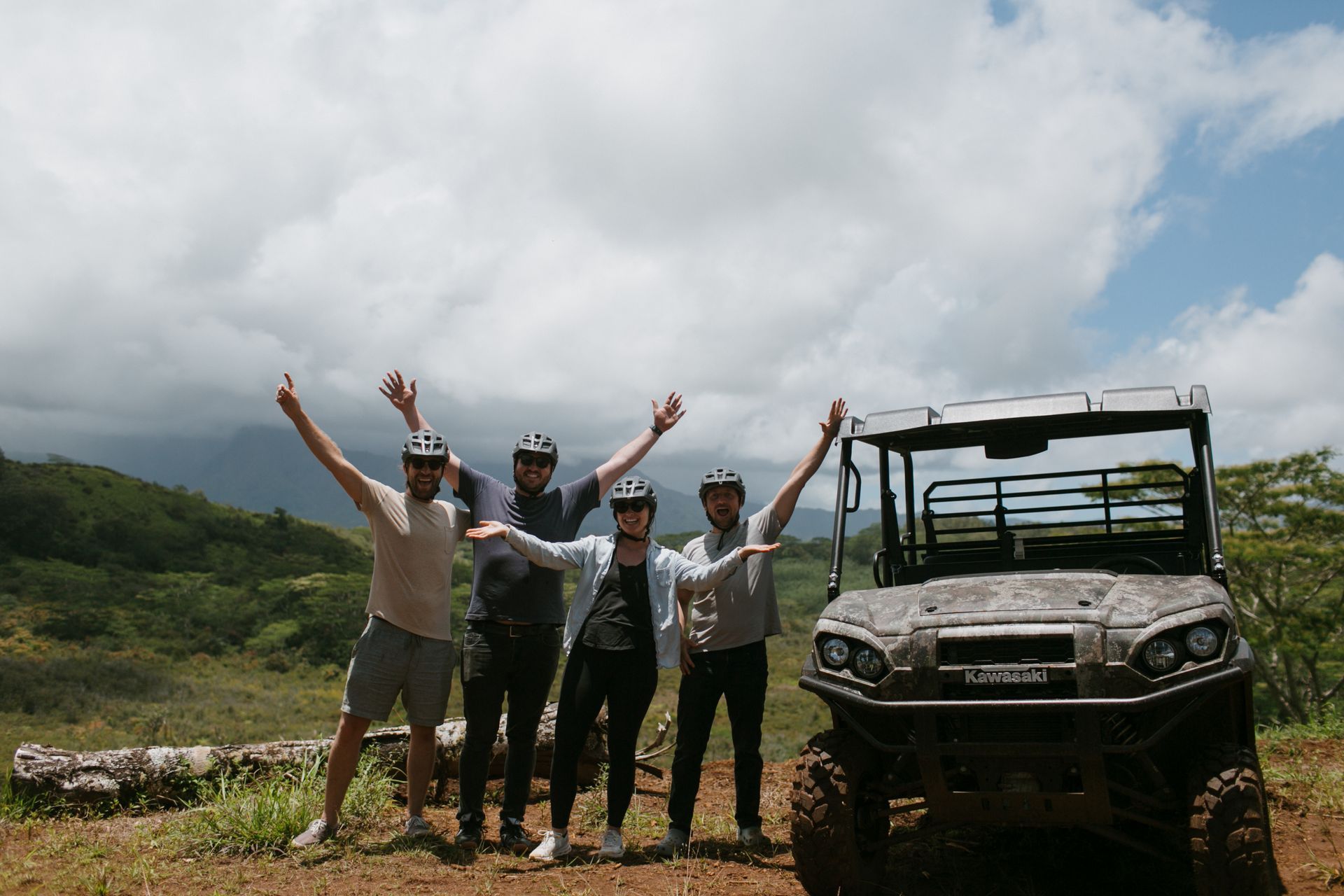 Four people raise their arms beside an off-road vehicle on a muddy trail under cloudy skies.