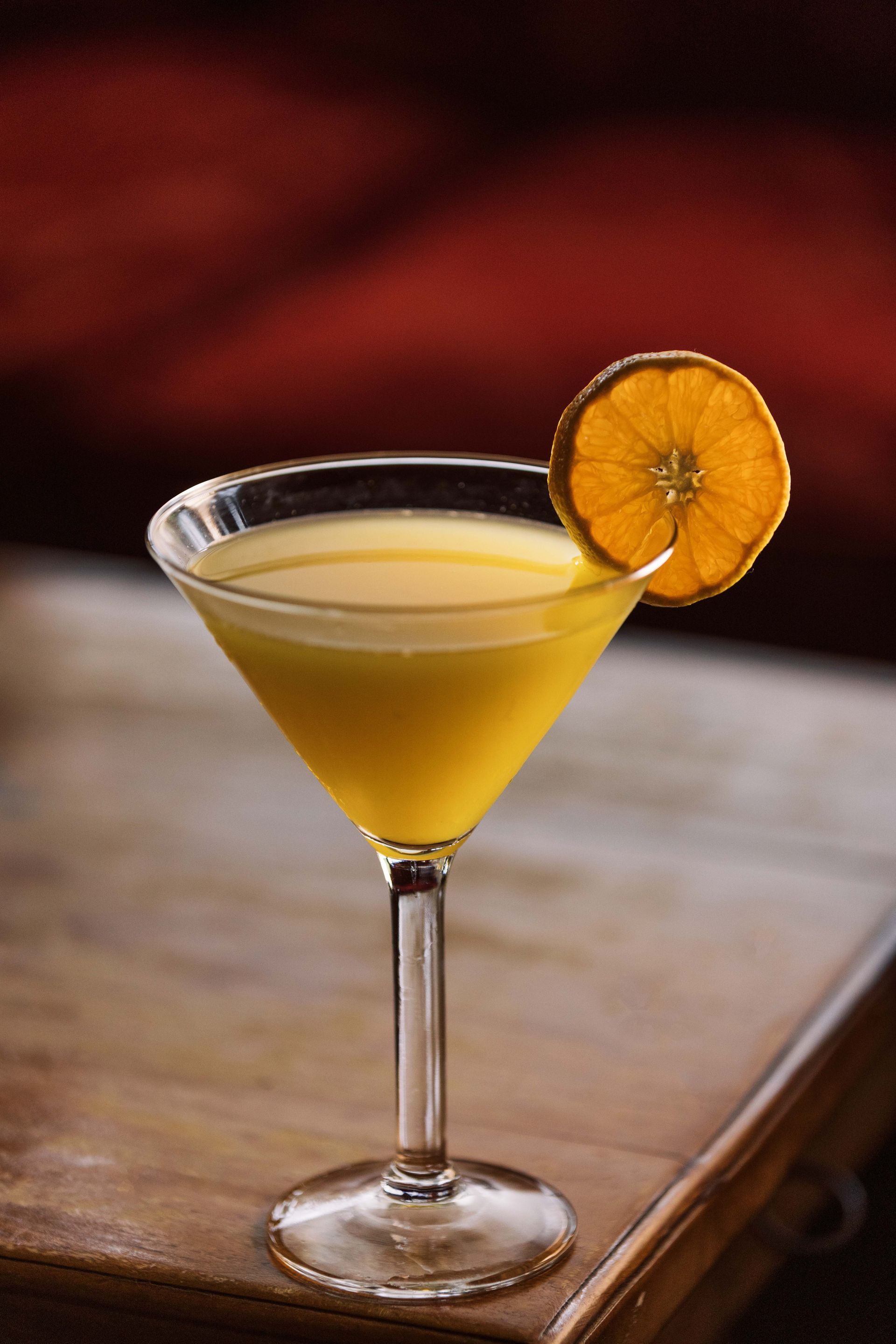 Martini glass with a yellow cocktail, garnished with a dried orange slice on a wooden table.