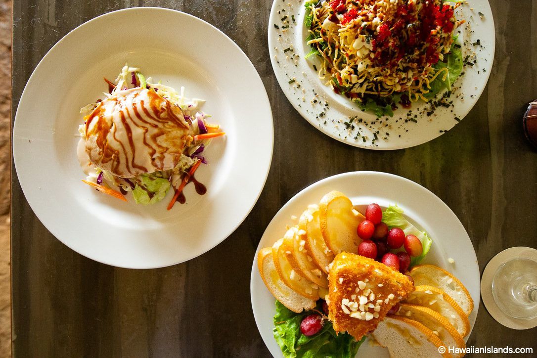 Three plated meals on a wooden table, including salad, pancakes, and a fruit dish with toast.