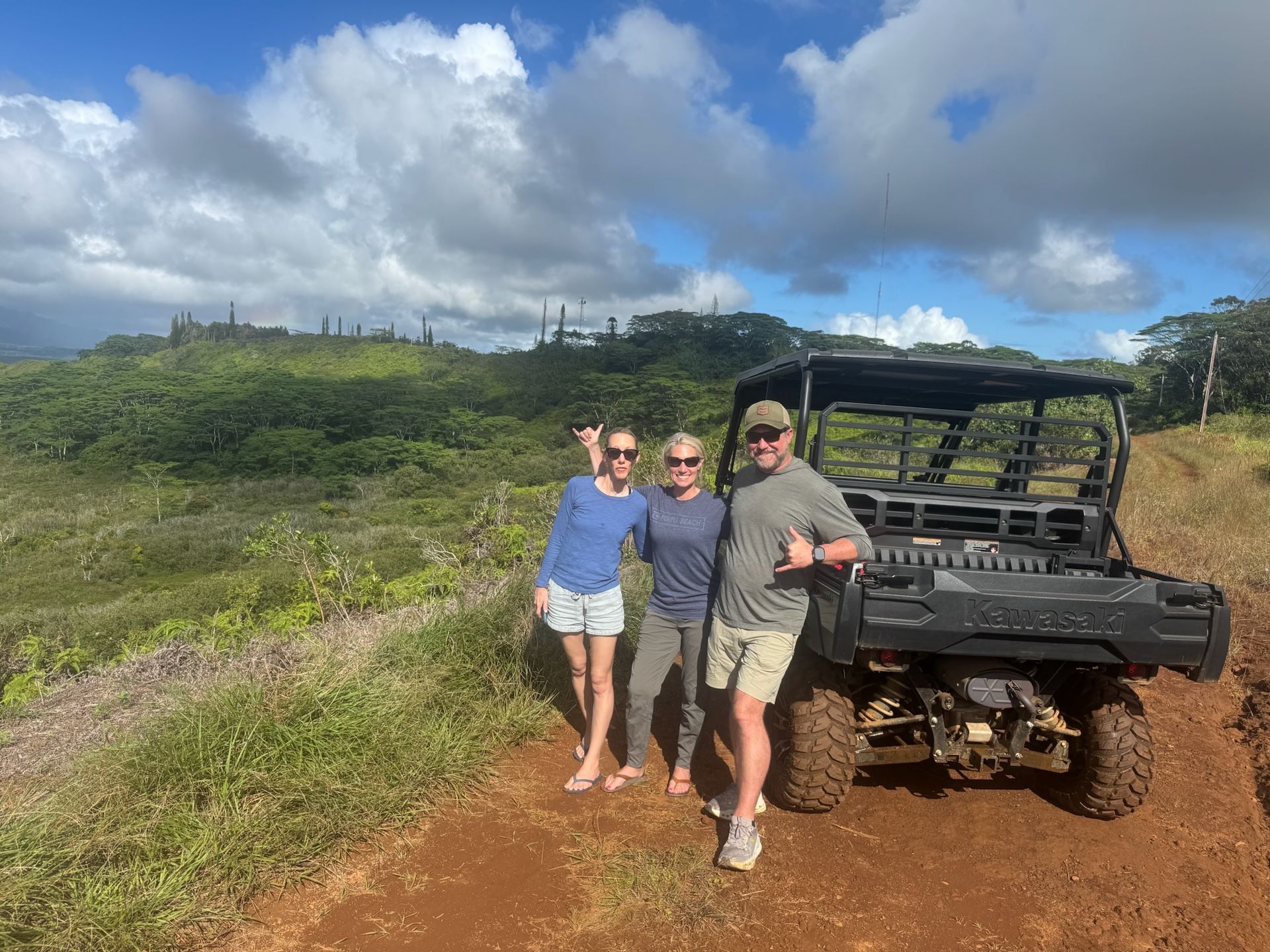 Three people posing beside a black off-road vehicle on a red dirt trail under a blue sky