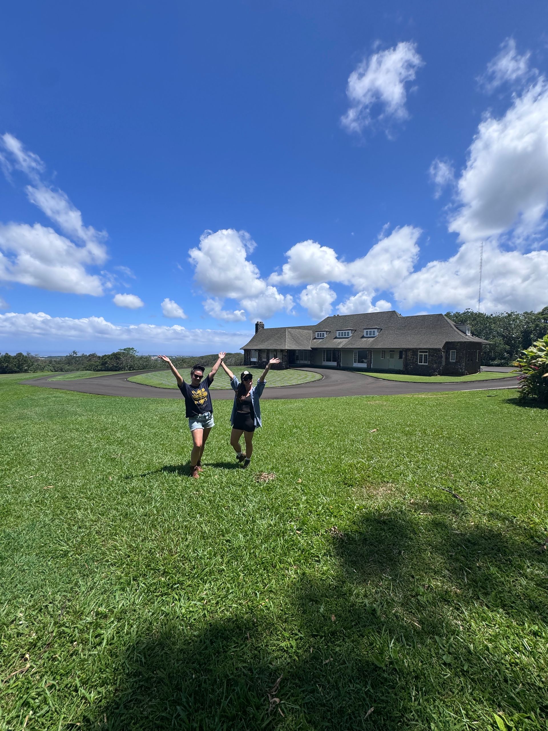 Two people standing in a green field under a bright blue sky near a large house