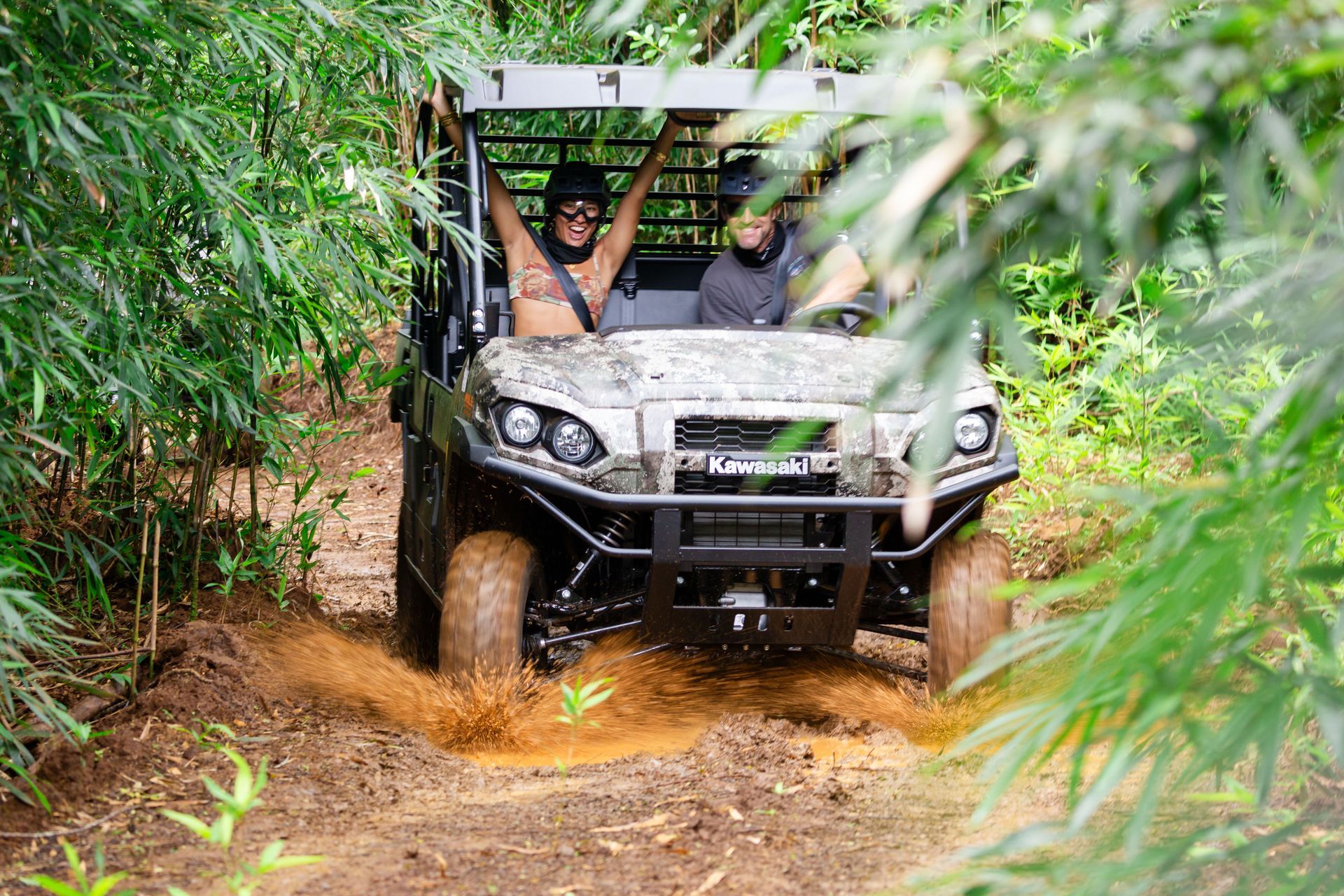 Off-road vehicle driving through muddy forest trail, with two passengers visible.