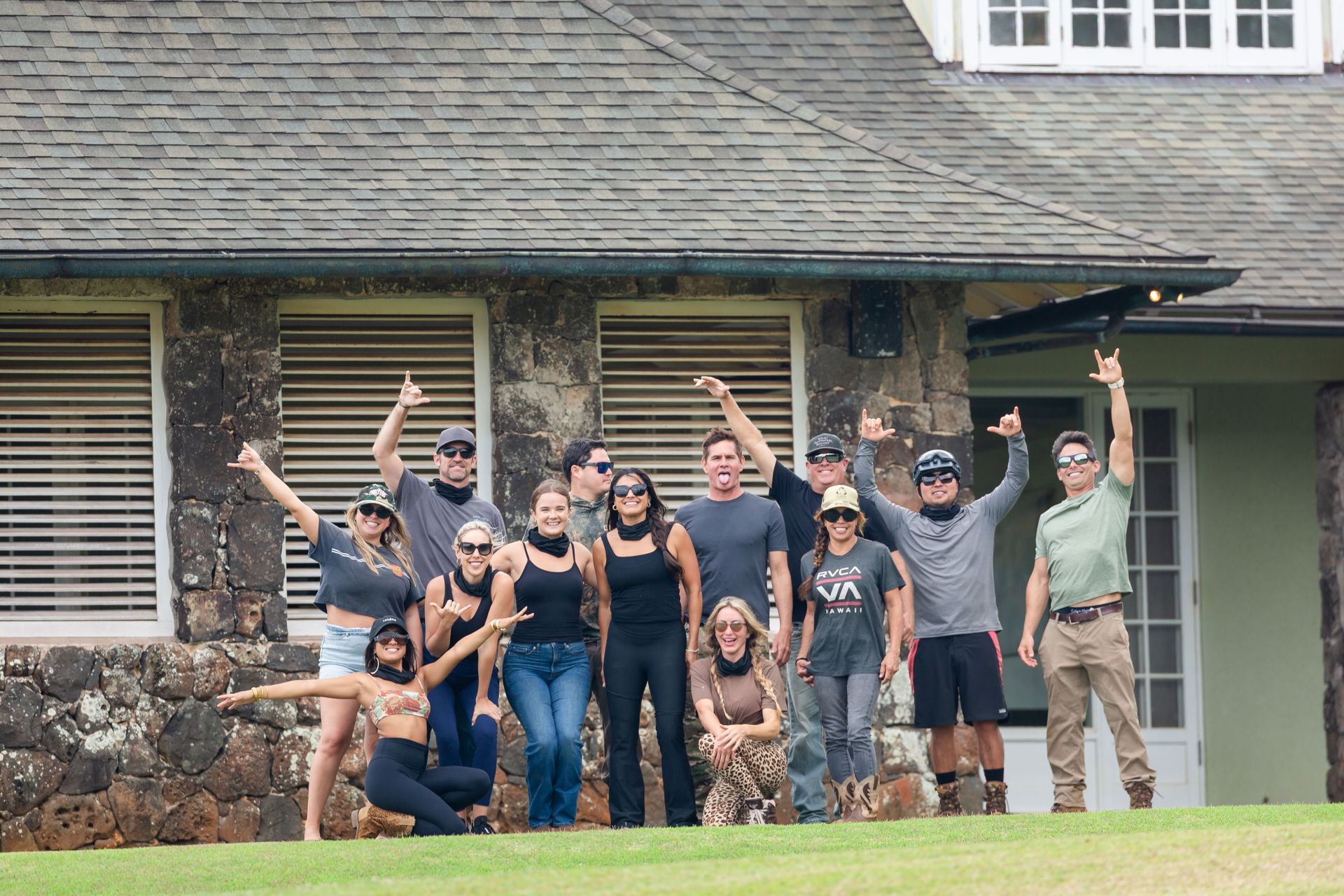 Group of people posing and raising arms in front of a stone house with green doors and lawn