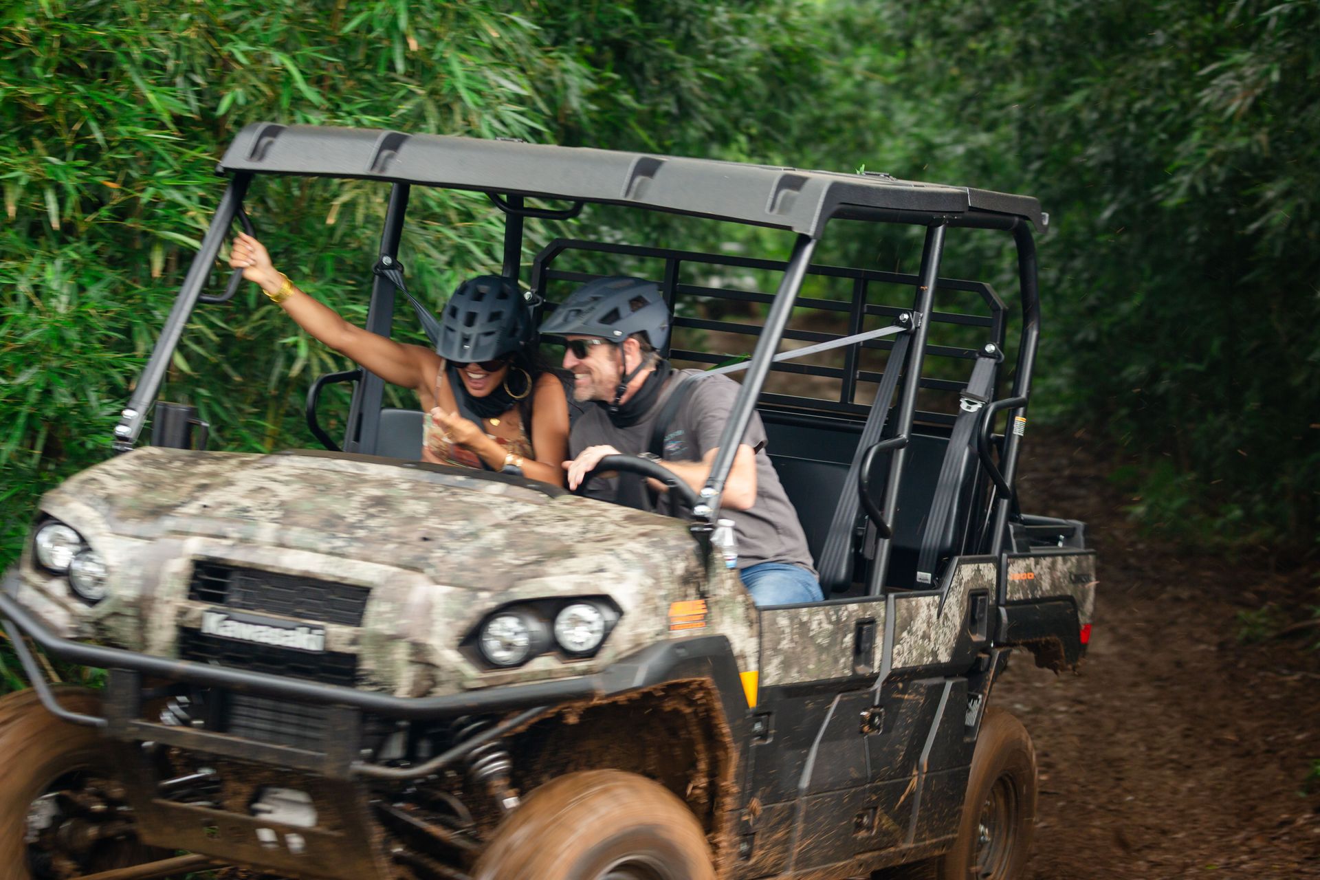 Two riders in a muddy off-road buggy driving through a dense green forest trail
