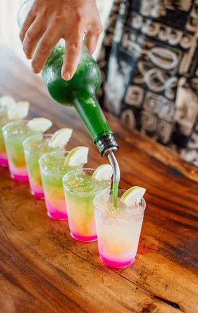 Hand pouring green drink into neon pink and lime shots on a wooden bar