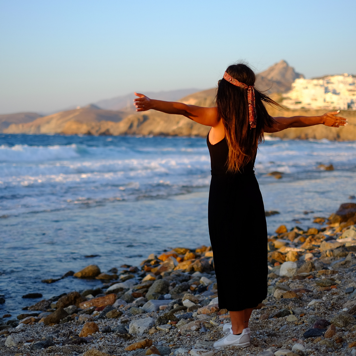 A woman in a black dress is standing on a rocky beach with her arms outstretched