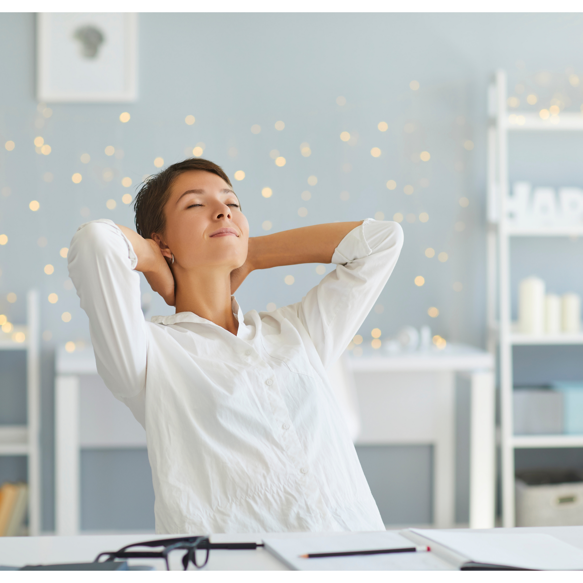 A woman is sitting at a desk with her hands behind her head.