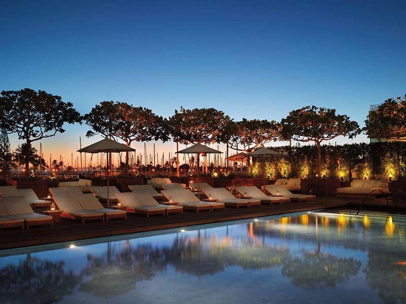 Poolside view at dusk with lounge chairs, trees, and blue sky.