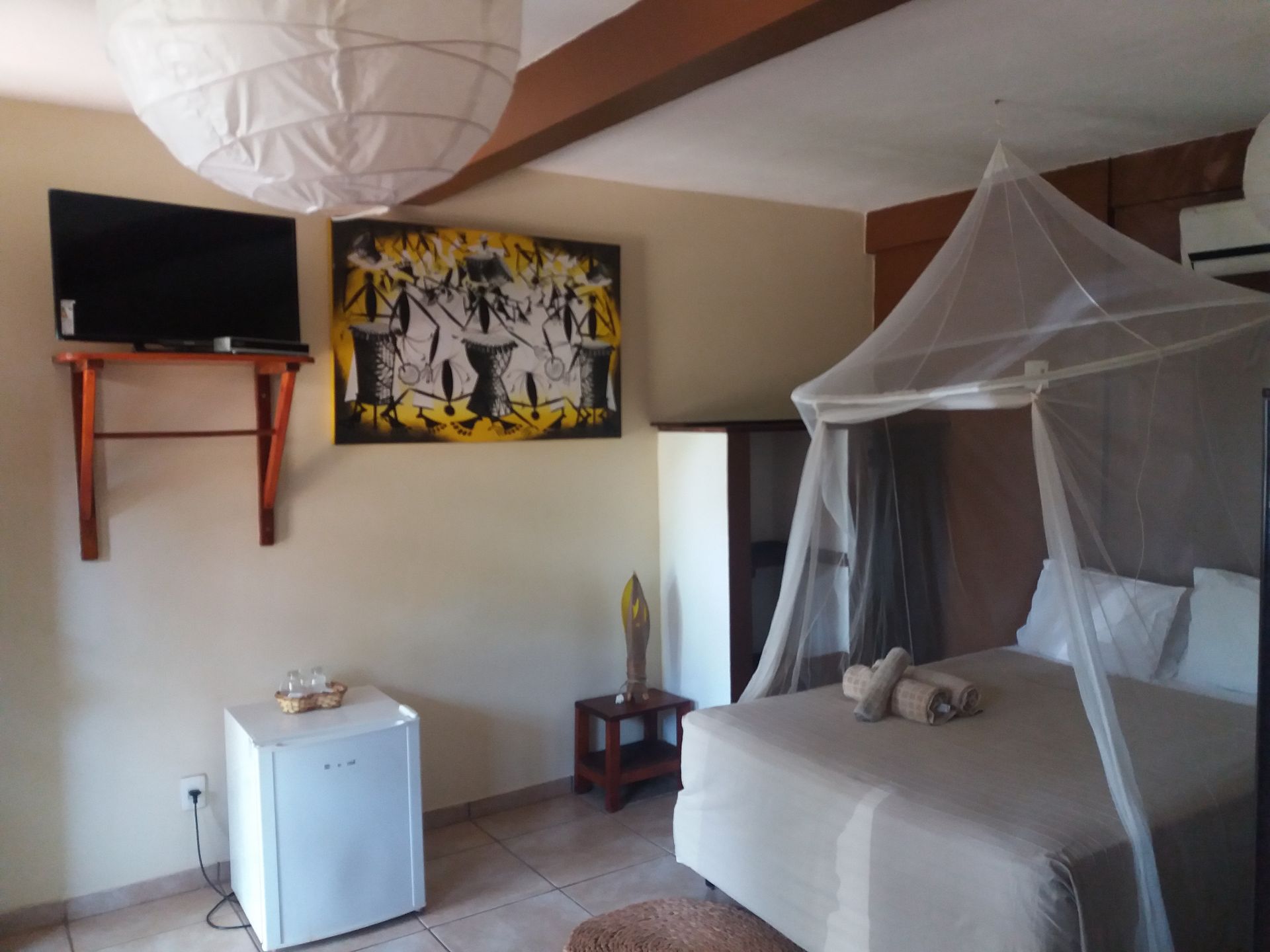 Bedroom with bed under mosquito net, TV, art, mini-fridge, and paper lantern.