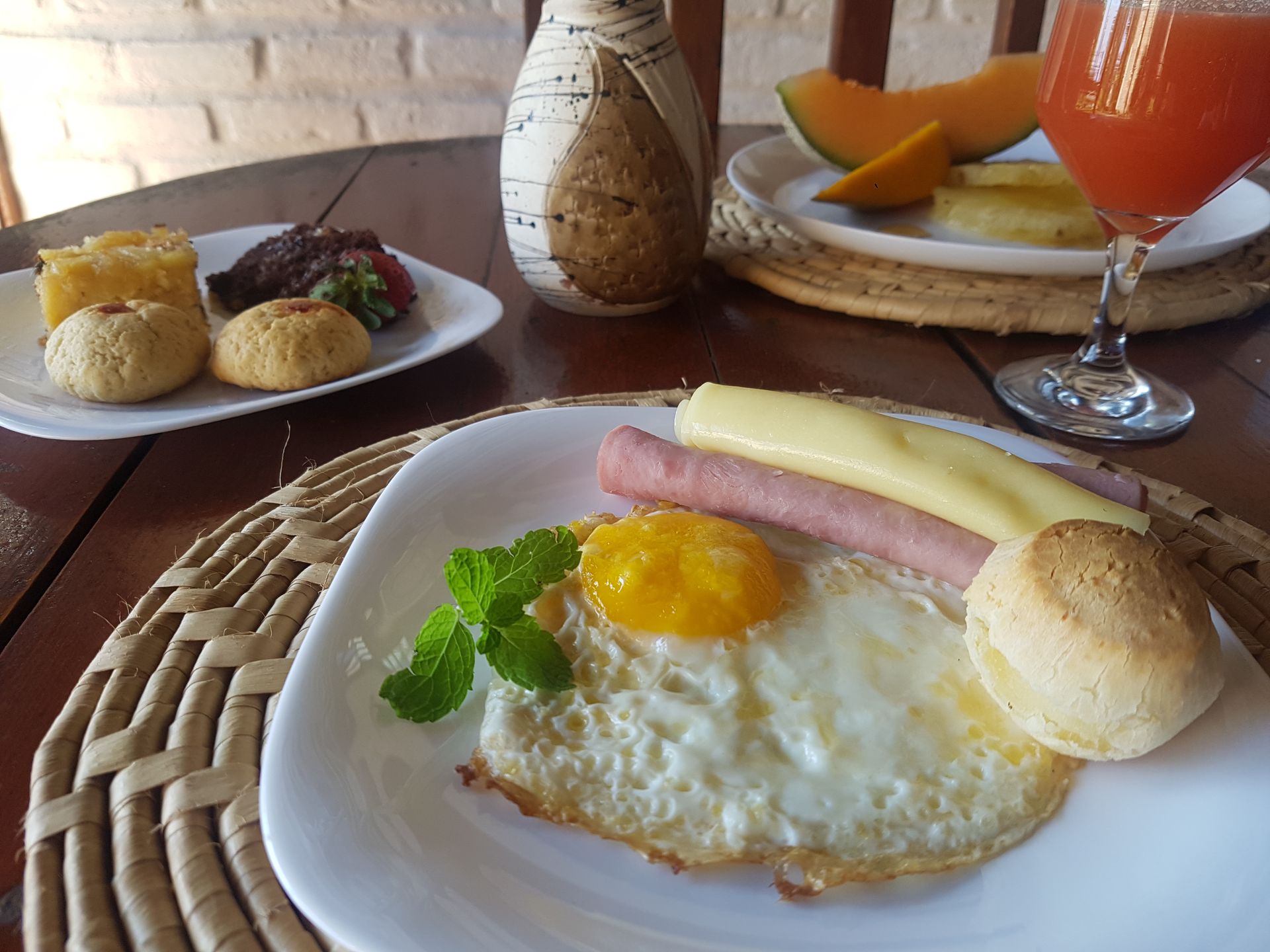 Breakfast plate with fried egg, sausage, cheese, bread, and juice. Fruit in background.