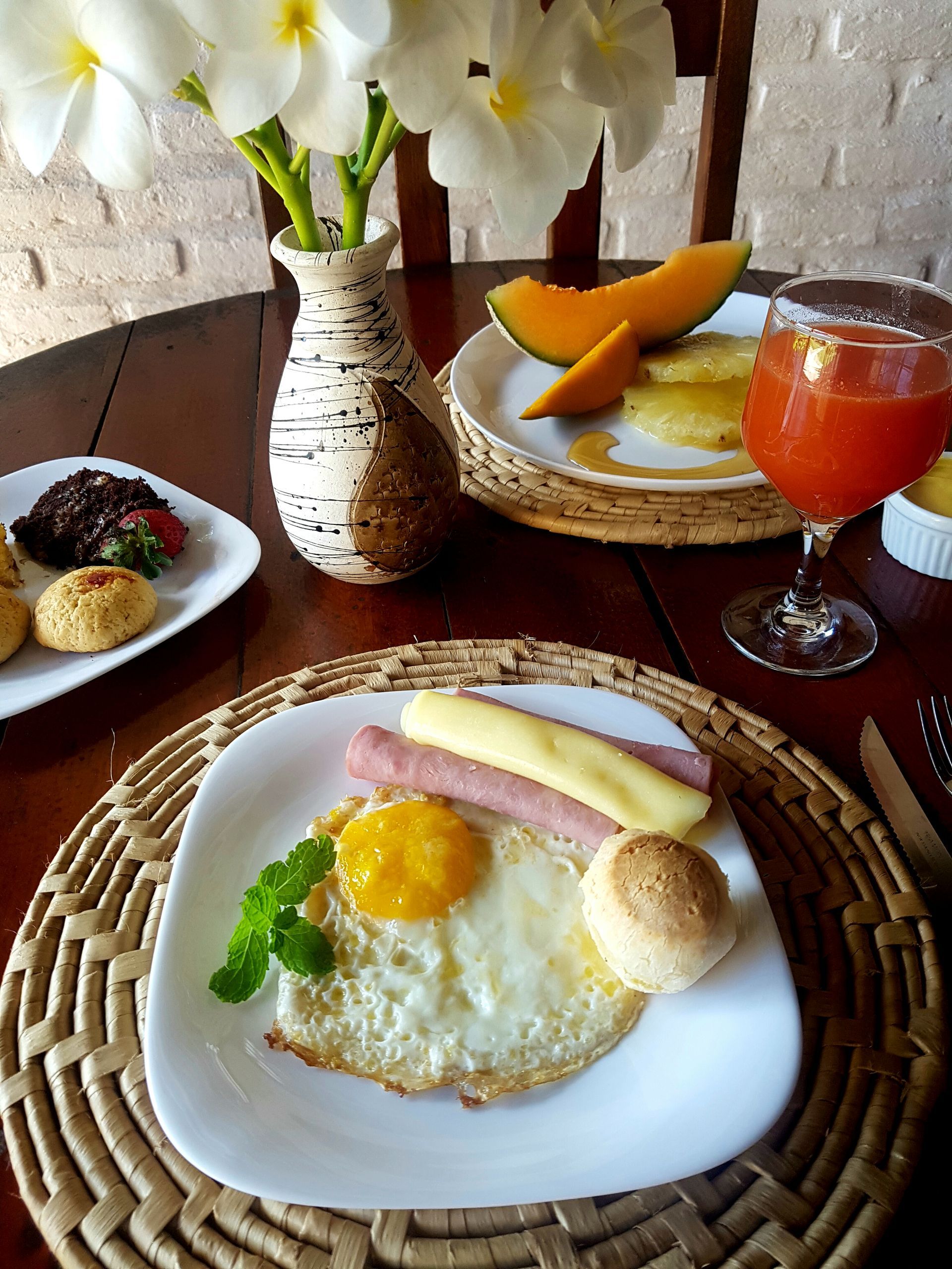Breakfast plate with fried egg, ham, cheese, and bread roll. Fruit and juice on the table, plus a vase of flowers.