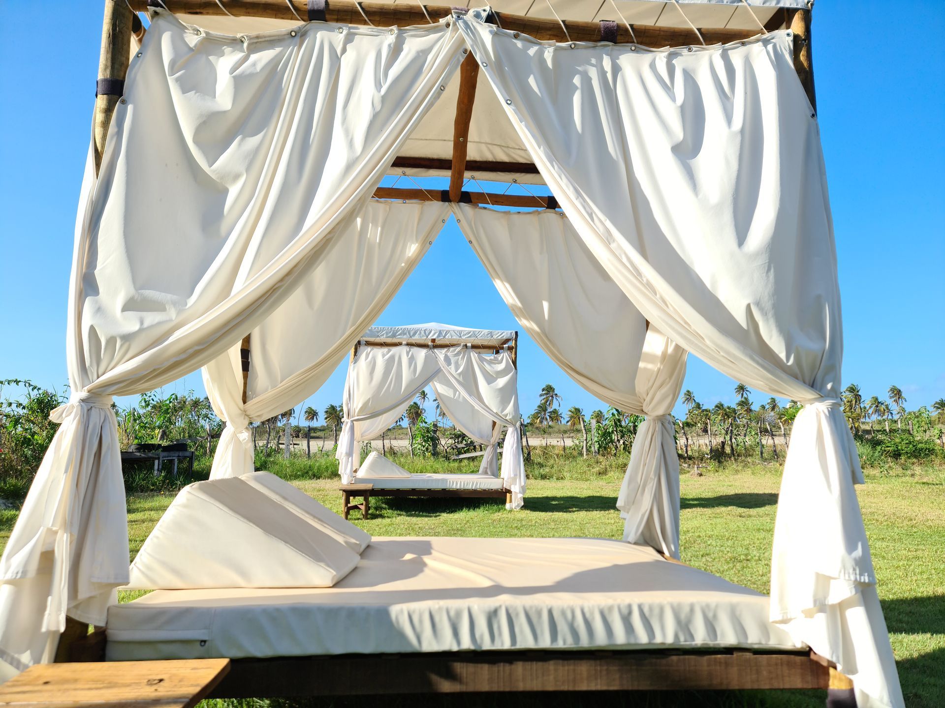 Outdoor canopied daybeds on grass with white curtains, blue sky, and another bed in the background.