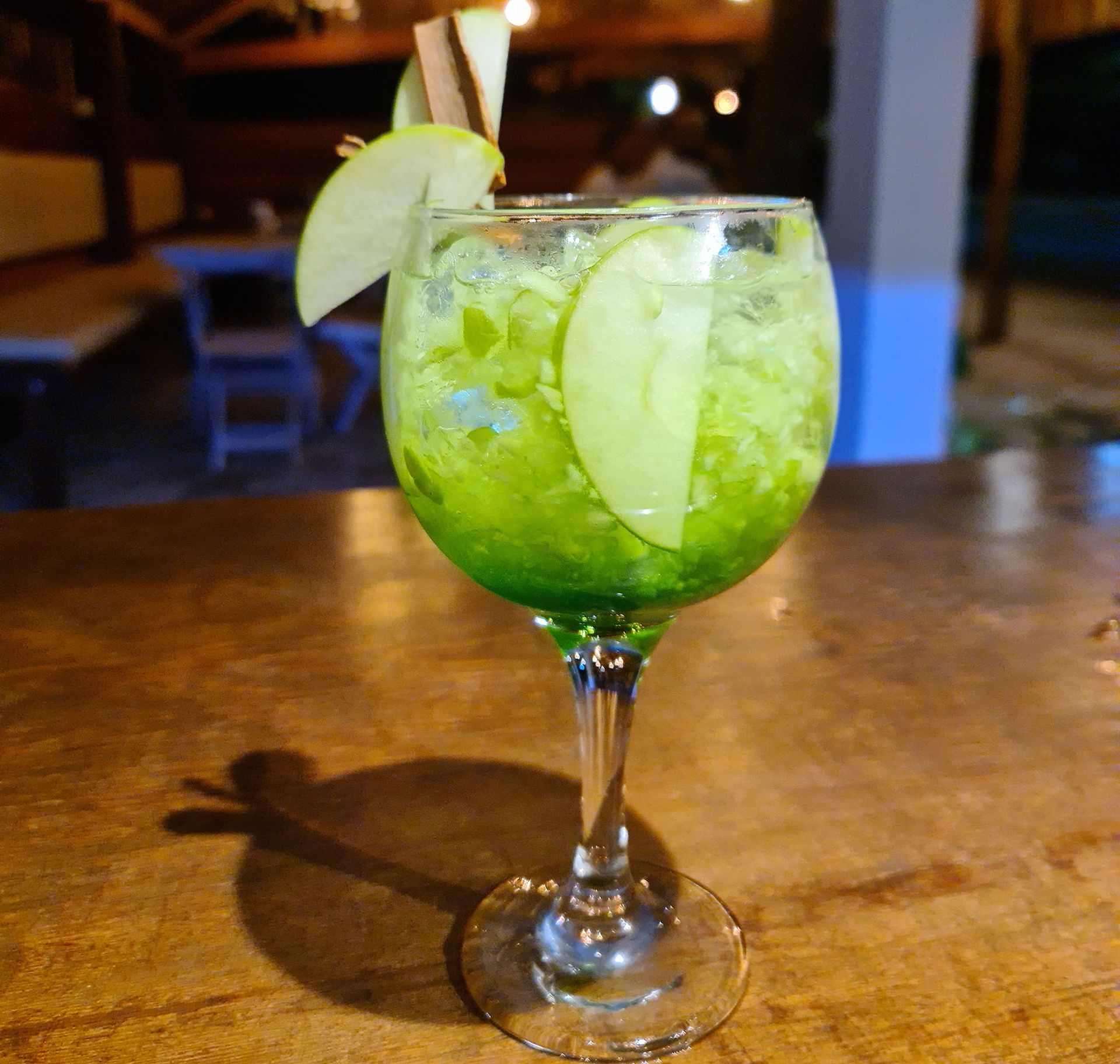 Green apple cocktail in a stemmed glass, garnished with apple slices on a wooden bar.