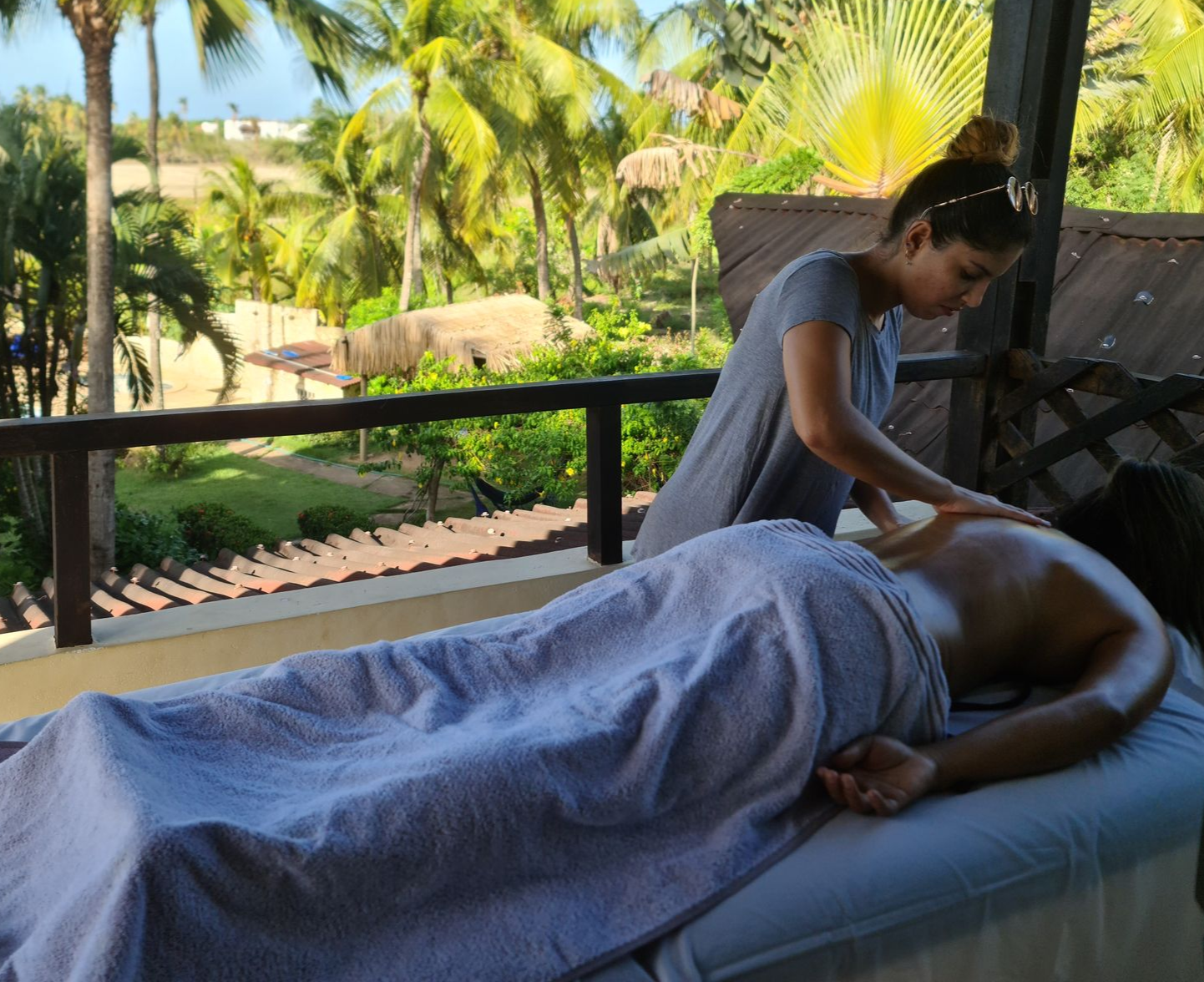 Woman giving a back massage on a covered outdoor patio overlooking lush greenery.