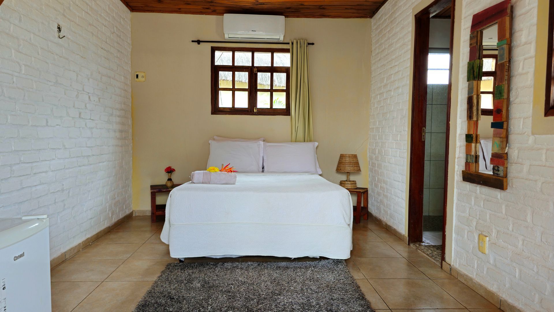 Bedroom with white brick walls, double bed, window, and rug. Door leads to bathroom.