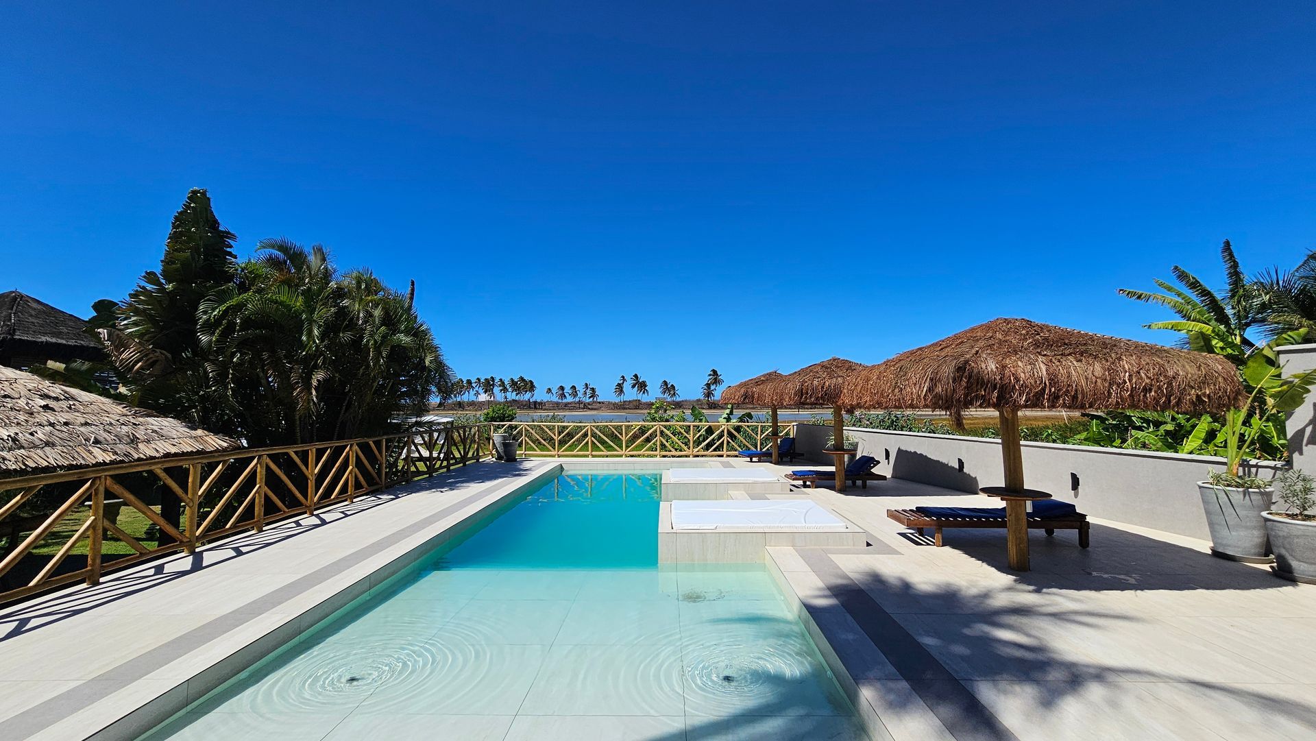 Pool with clear water and thatched umbrellas under a bright blue sky.