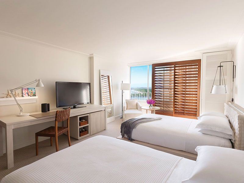 Hotel room with two twin beds, desk, TV, and open sliding doors to a sunny view.