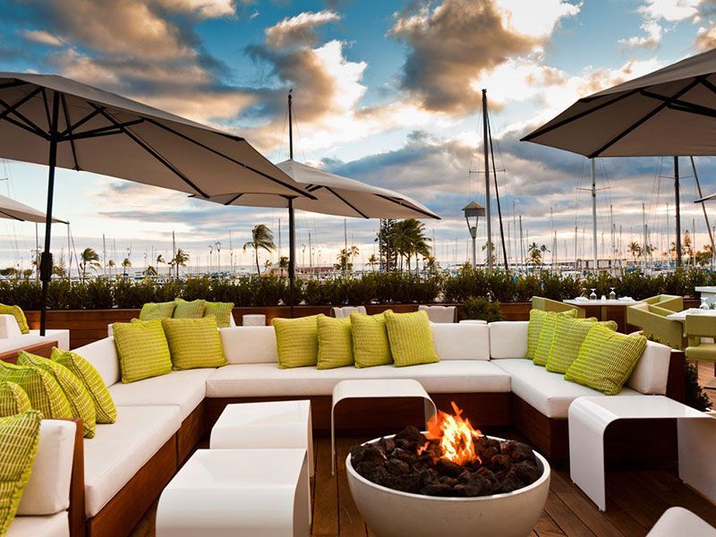 Outdoor lounge area with white couches, green pillows, fire pit, umbrellas, and harbor view.