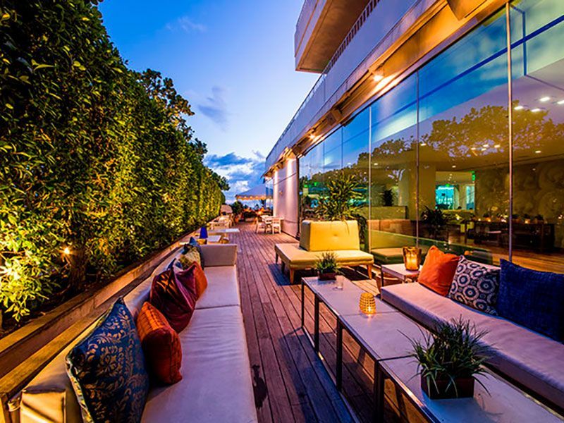 Rooftop lounge at dusk with plush couches, colorful pillows, lush greenery, and illuminated glass walls.