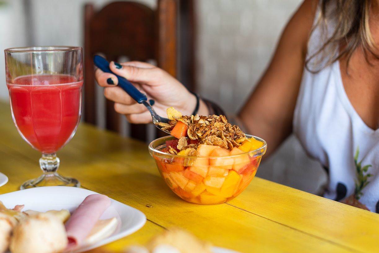 Woman eating fruit salad with granola, red juice, breakfast plate on yellow table.