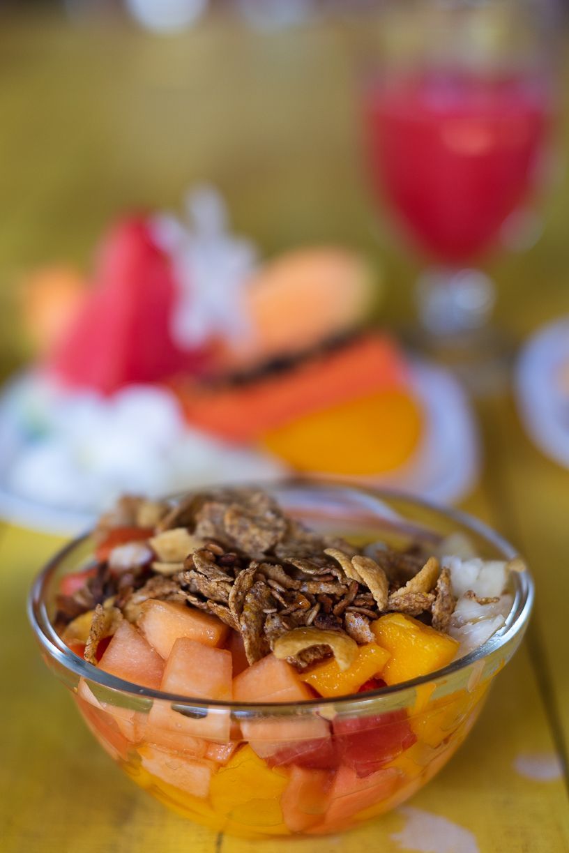 Fruit salad in glass bowl topped with granola; sliced fruit and red drink in background.