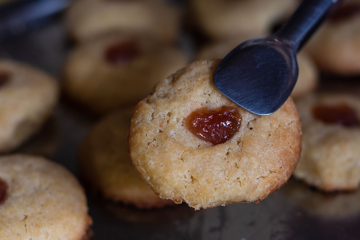A hand holding a cookie with tongs; others visible on a tray, each with a jam center.