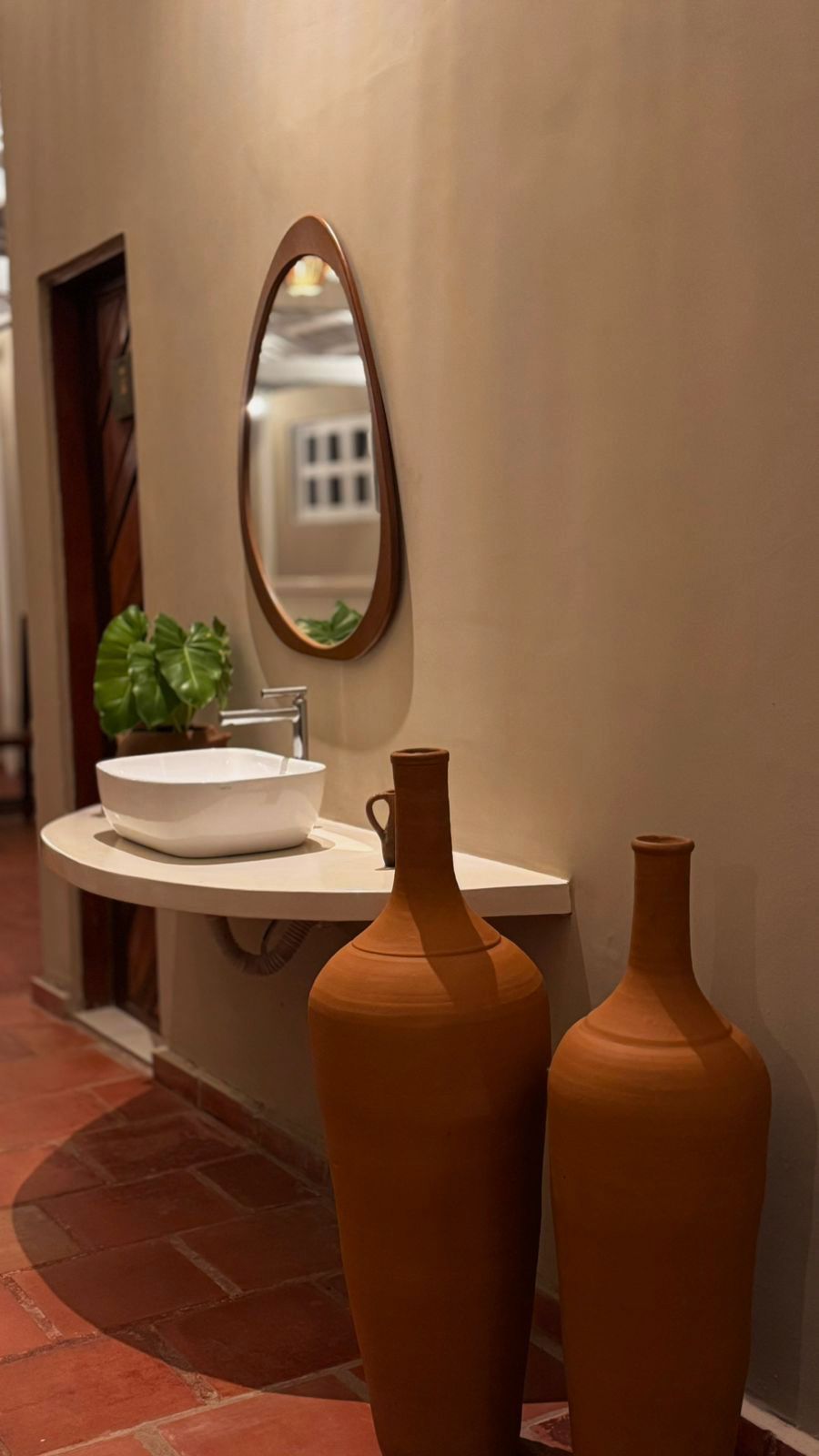 Bathroom with white sink, brown terracotta vessels, oval mirror, and red tile flooring.