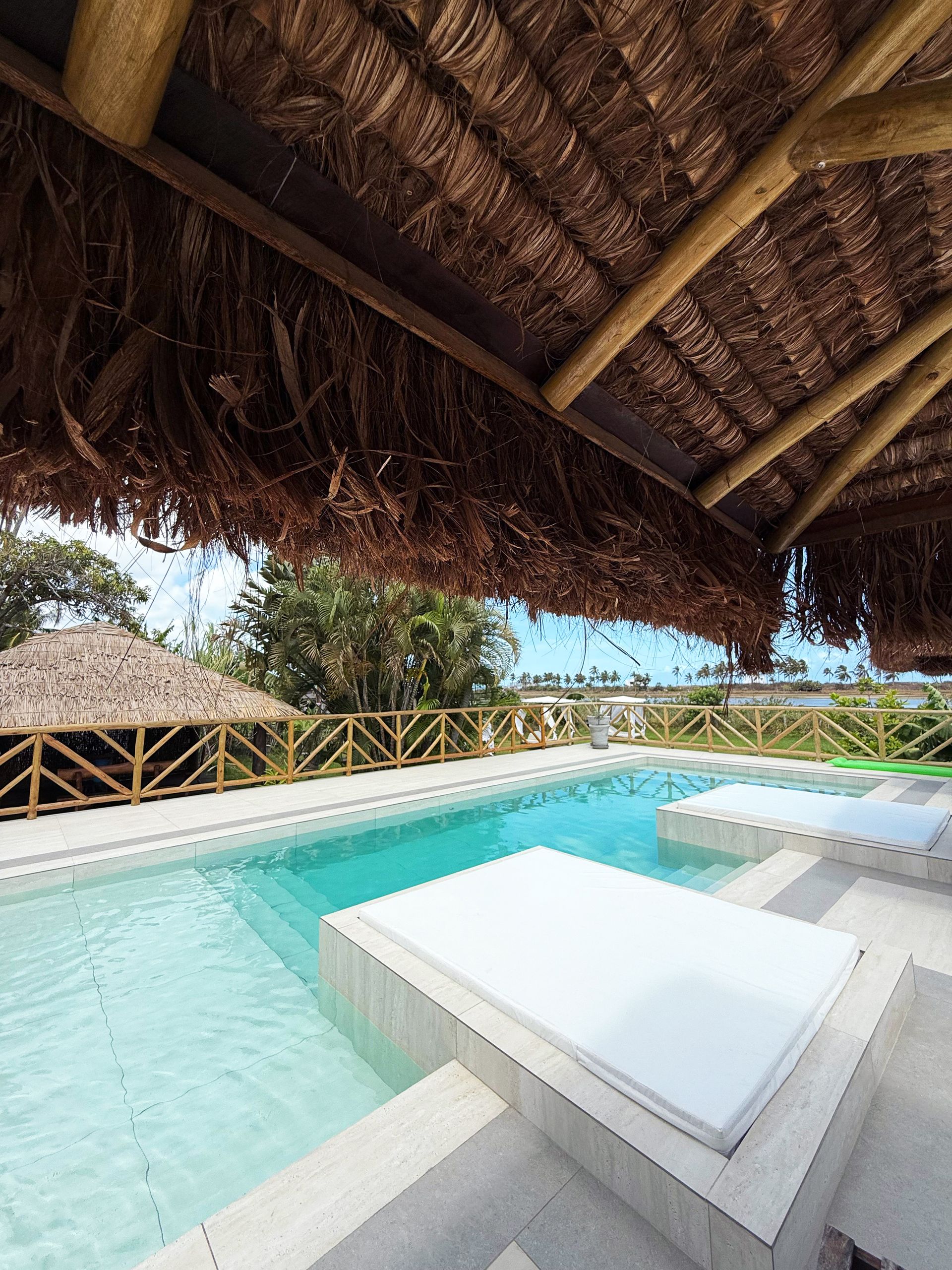 Pool with clear water under a thatched roof structure, adjacent to a wooden railing and lush greenery.
