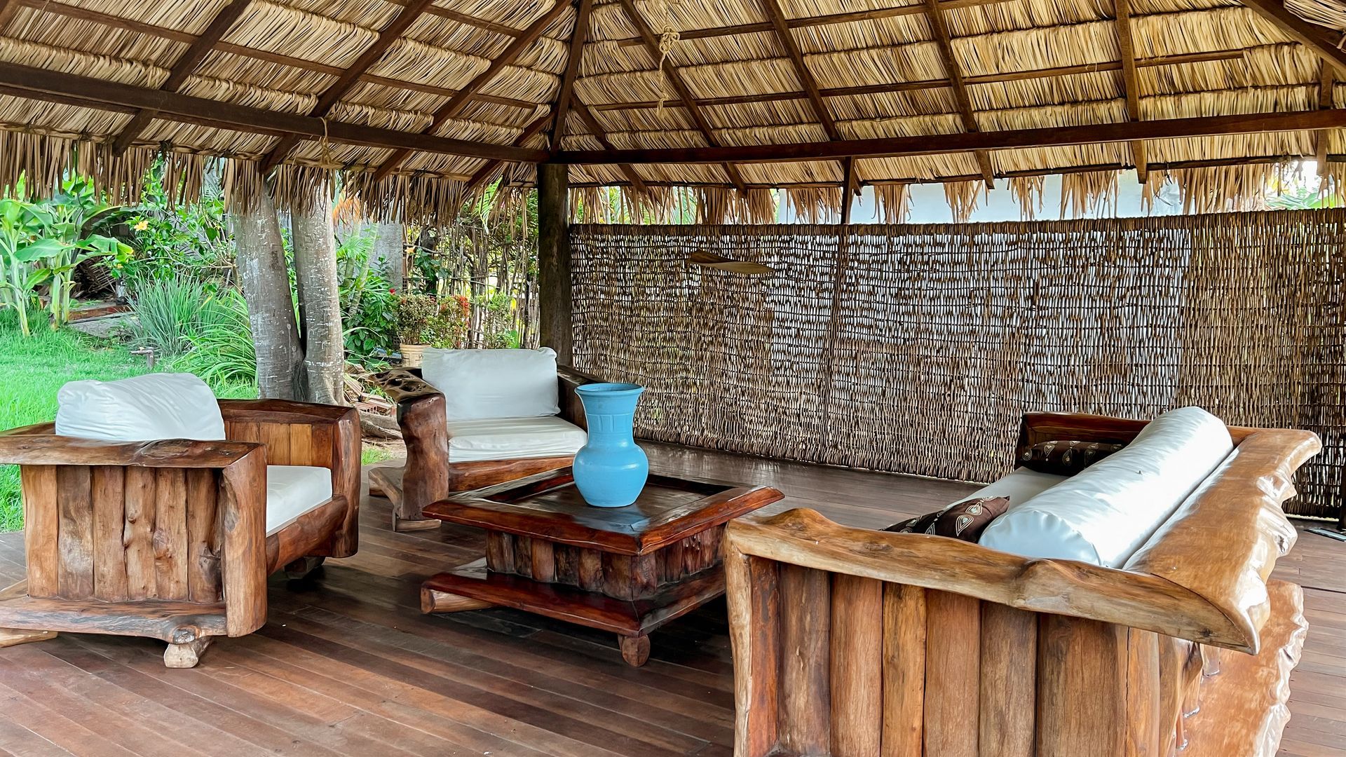 Wooden outdoor seating area with a thatched roof, surrounded by greenery.
