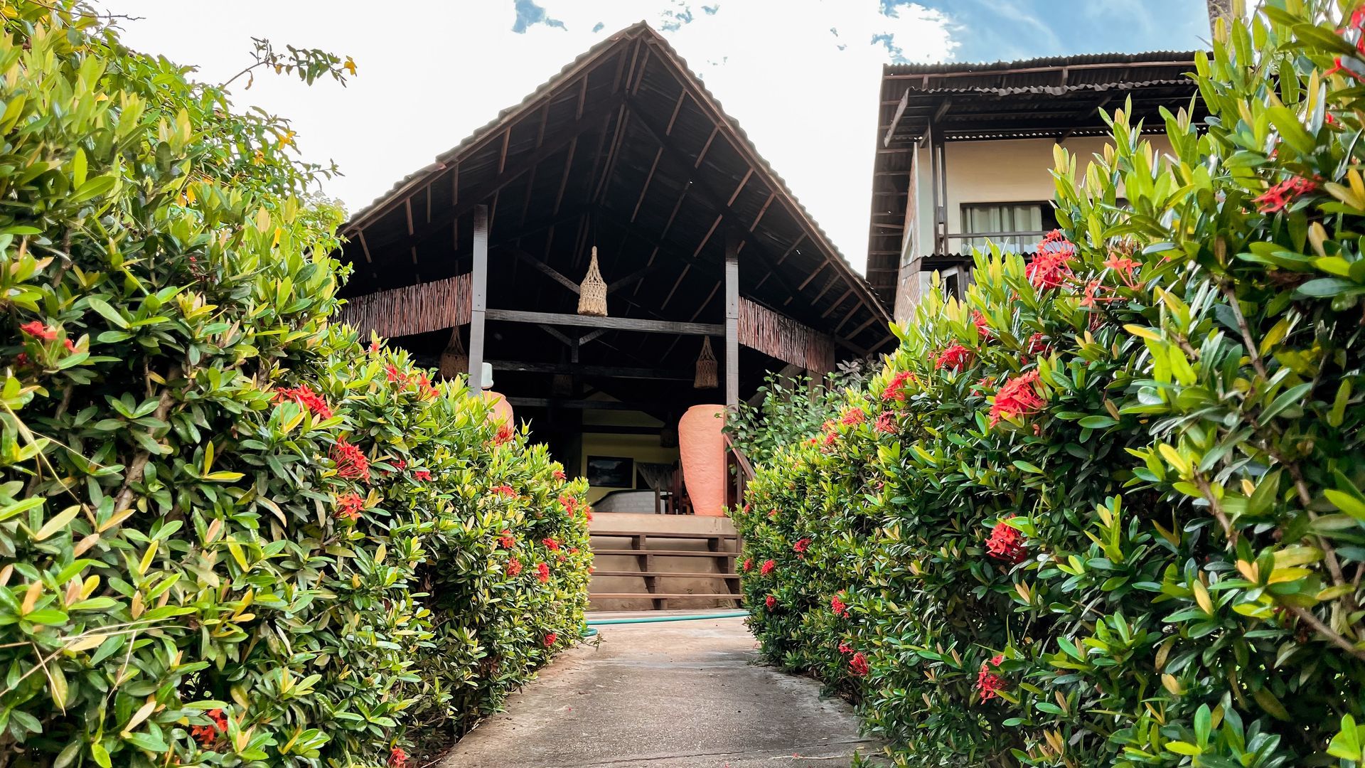 Path through manicured hedges leads to a thatched-roof structure. Red flowers dot the green bushes.