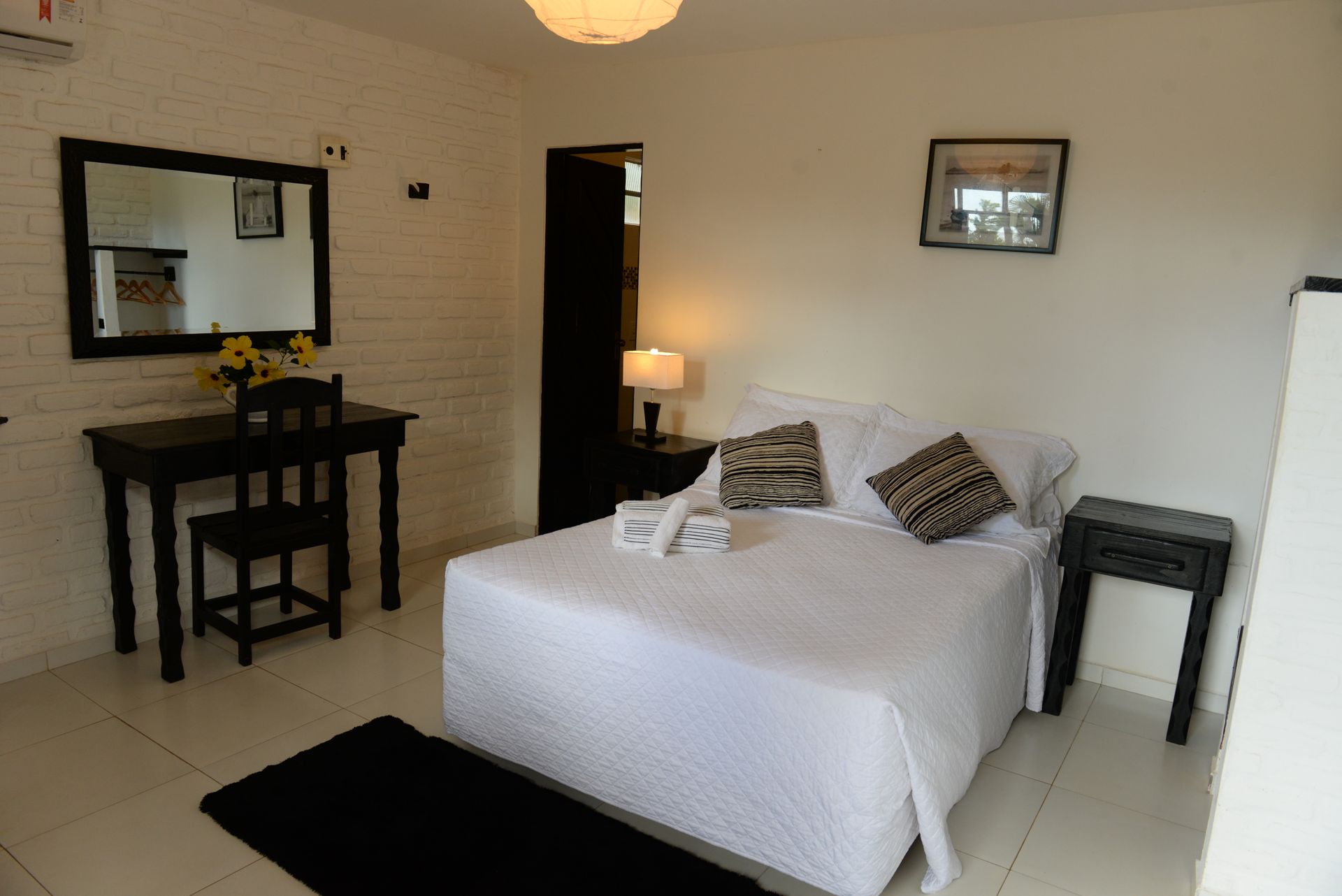 Bedroom with a bed, desk with mirror, side table, and black rug on white tiled floor.
