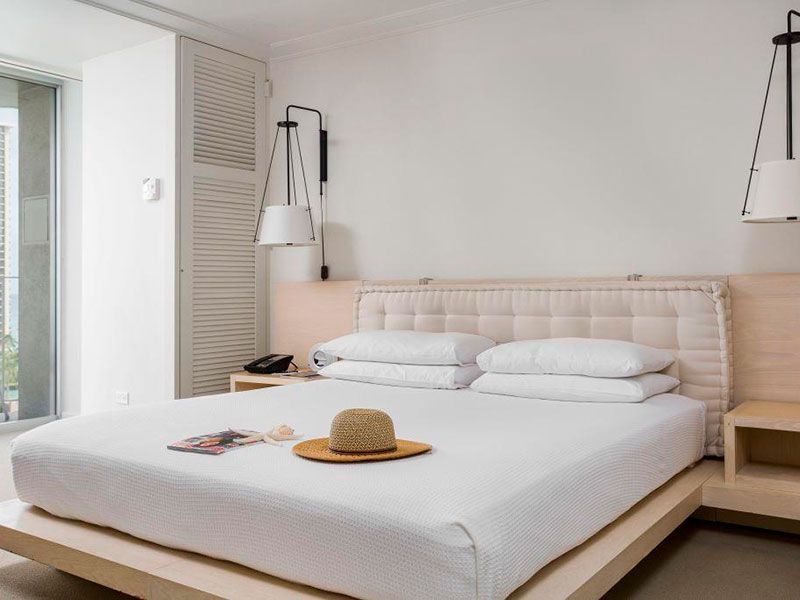White hotel bedroom with bed, nightstands, lamps, and a straw hat.