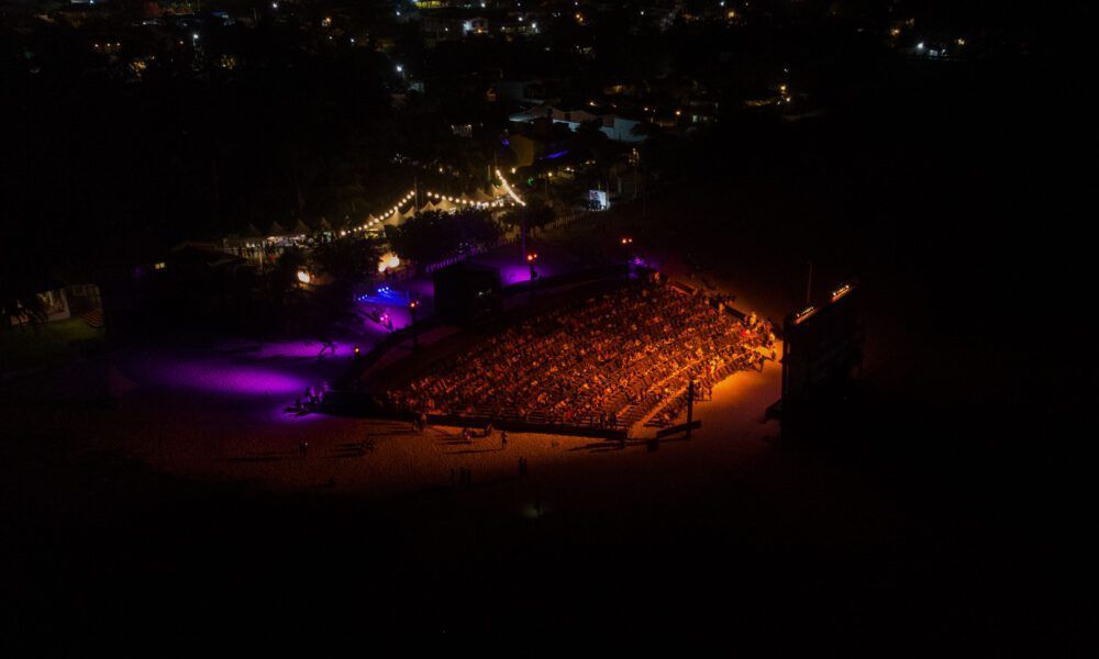 Nighttime outdoor concert with a large crowd and stage lit with purple and orange lights.