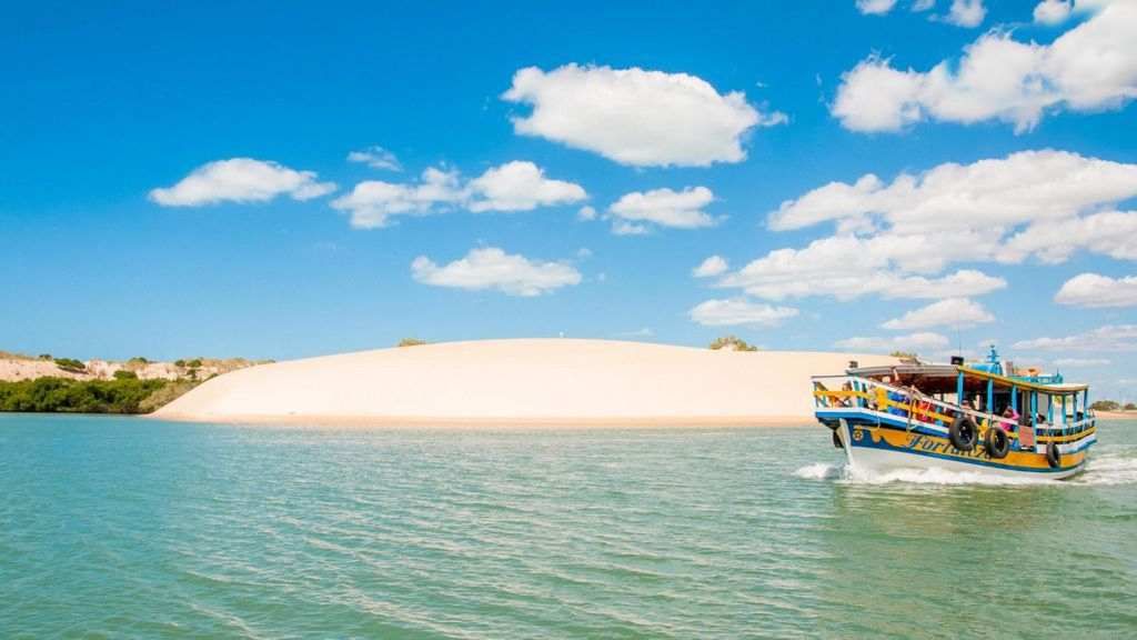 Boat on blue water with sand dunes and bright blue sky.