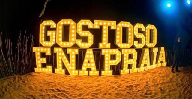 "GOSTOSO NA PRAIA" illuminated sign on a sandy beach at night.
