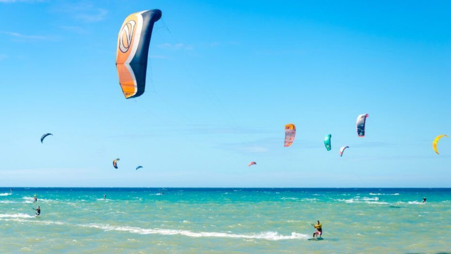 Kitesurfers on turquoise water under a blue sky, colorful kites flying.