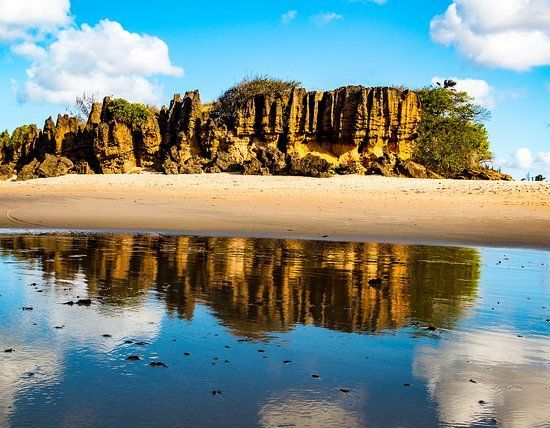 Beach with eroded sandstone cliffs and their reflection in the wet sand under a blue sky.