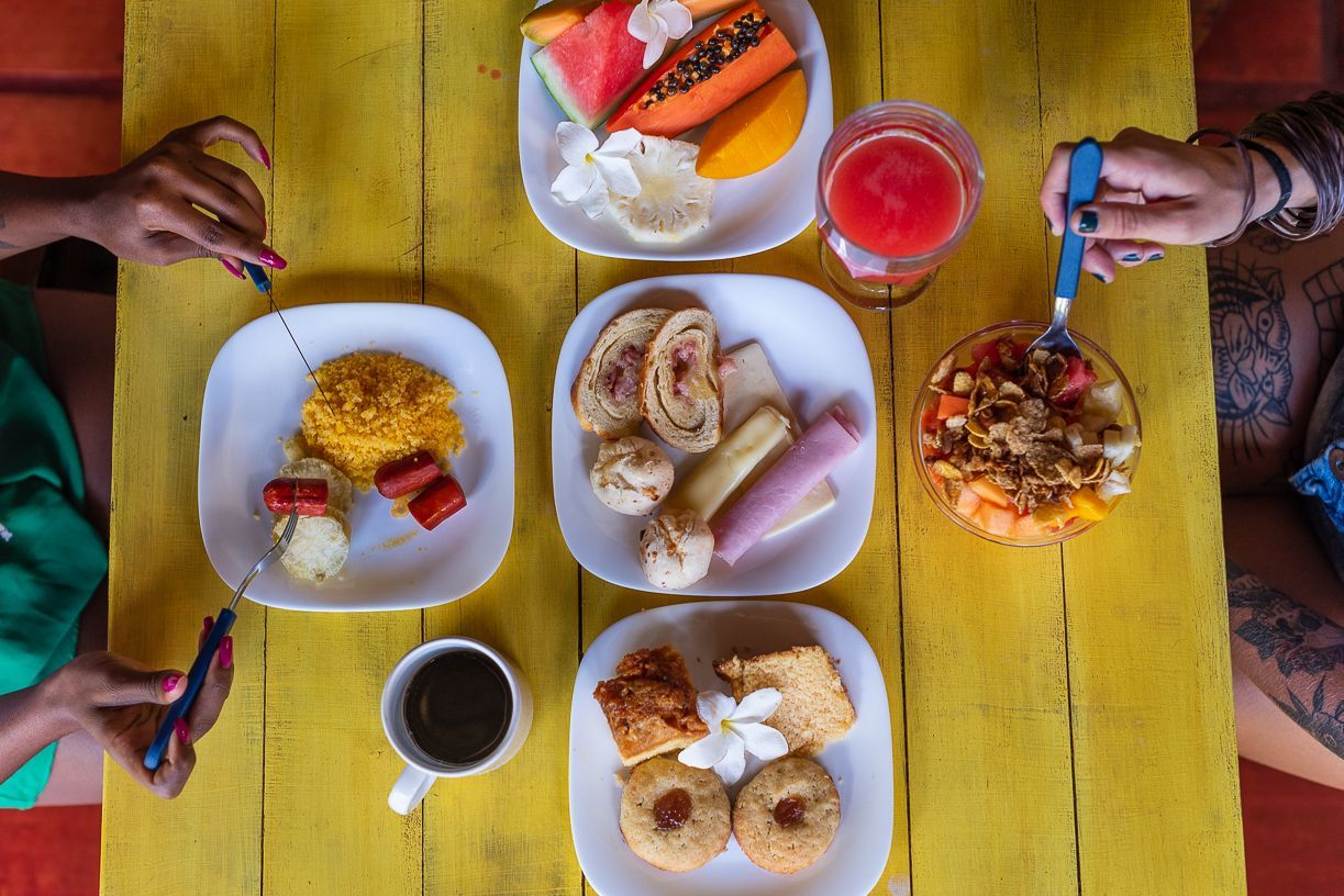 Overhead view of a yellow table with five plates of breakfast food, two hands holding utensils.