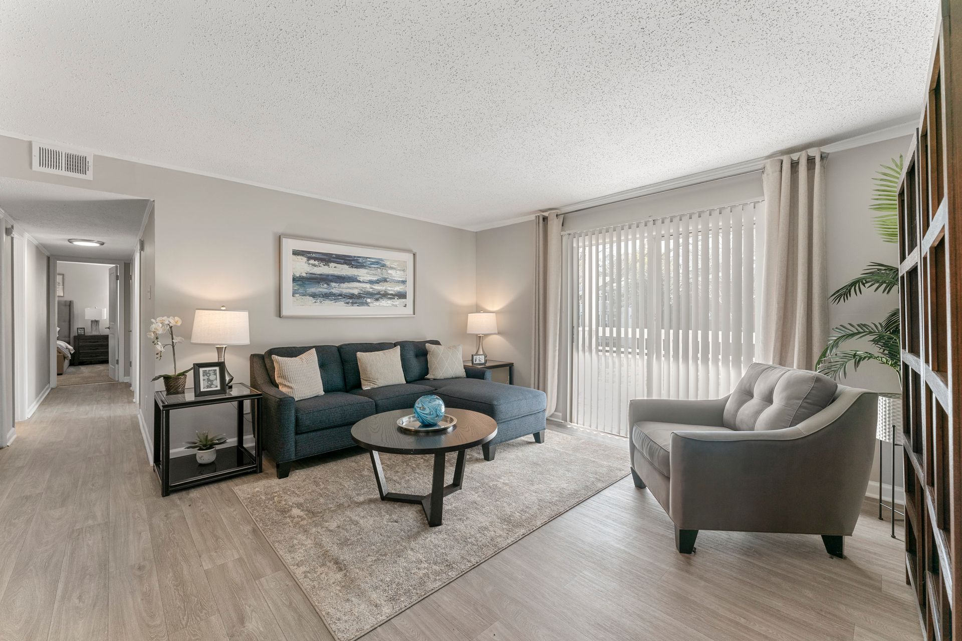 Bright living room with teal sofa, gray chairs, large window blinds, and a glass coffee table.