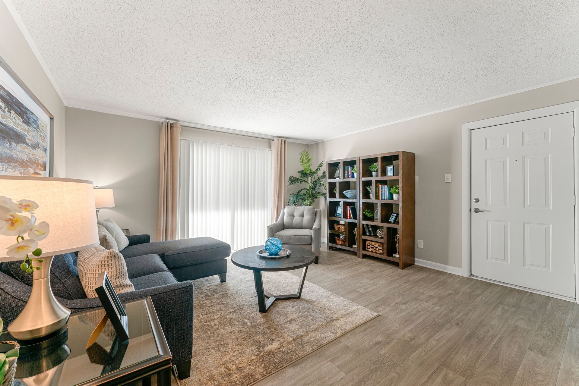 Bright living room with gray sofa, round coffee table, bookcase, and large window with curtains.